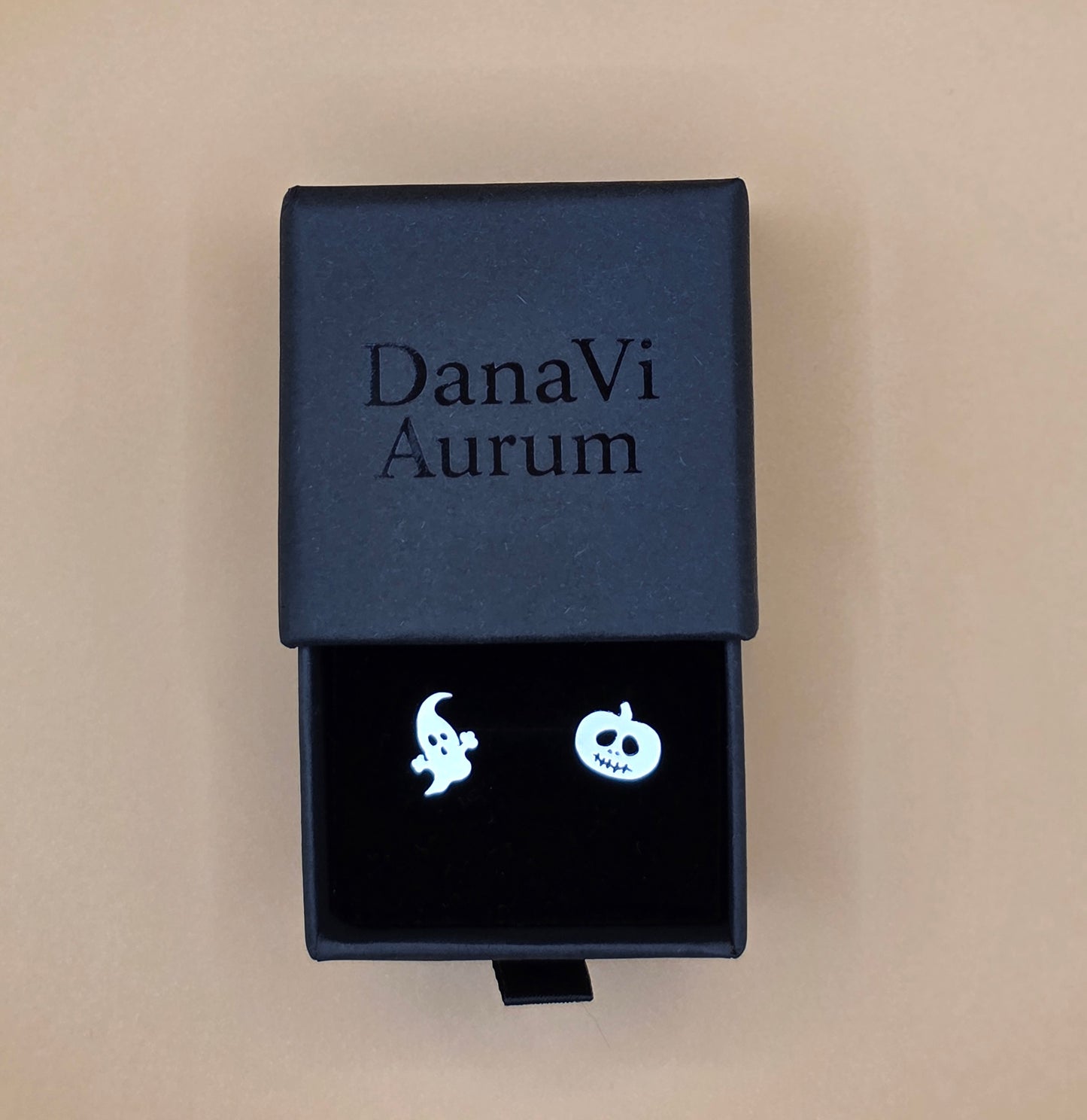 Black jewelry box with 'DanaVi Aurum' branding containing ghost-shaped earrings on a beige background.