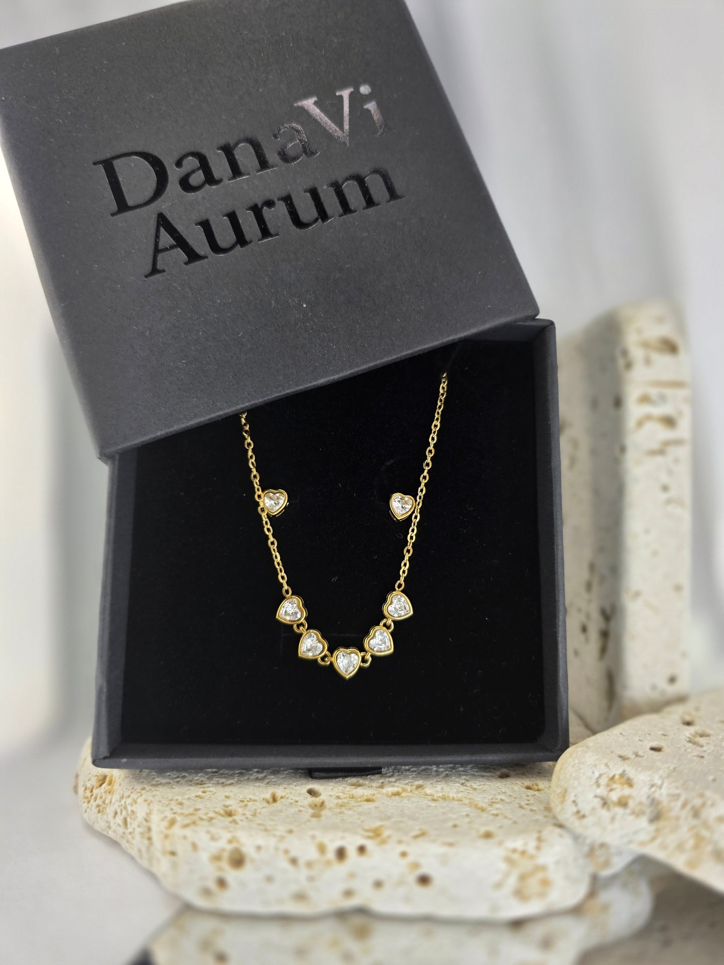 Gold necklace with heart-shaped pendants in a DanaVi Aurum box on a textured surface.