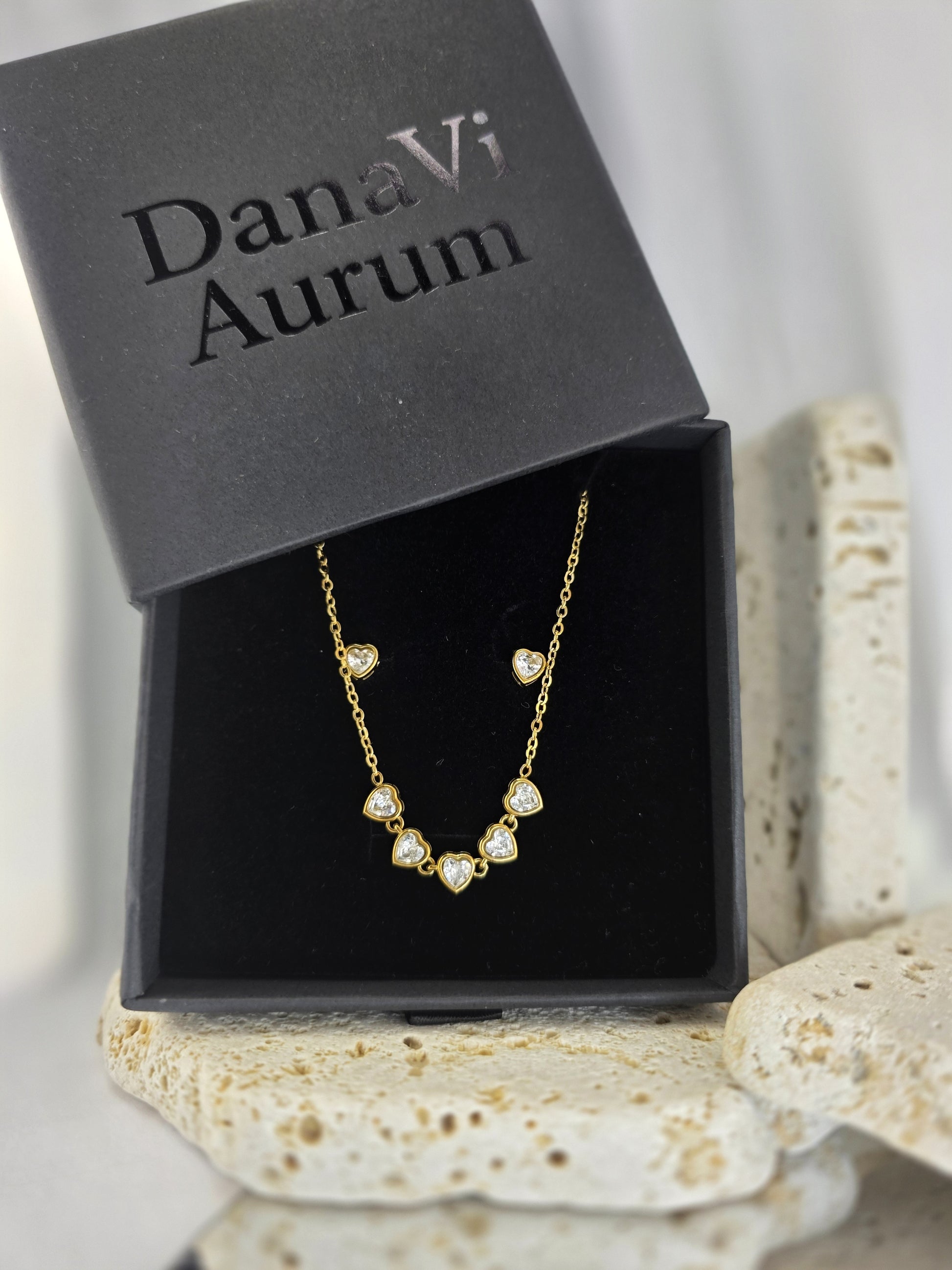 Gold necklace with heart-shaped pendants in a DanaVi Aurum box on a textured surface.